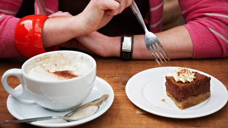 A lady's arms with a red cardigan and chunky red bracelet rests on a wooden table. A fork is held in their hand, poised over the top of a slice of cake on a white place, next to a large mug of frothy white coffee.